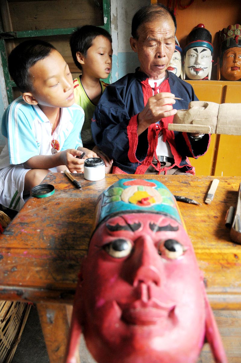 Le 18 août 2013, dans le district autonome Maonan du Huanjiang dans la province du Guangxi, Tan Shengci enseigne à deux garçons, comment sculter un masque de la danse Nuo. (Photo : Xinhua)